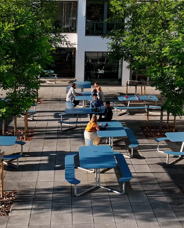 Seks blå Plateau Picnic I borde i en gård med store grønne træer, her sidder fire personer ned