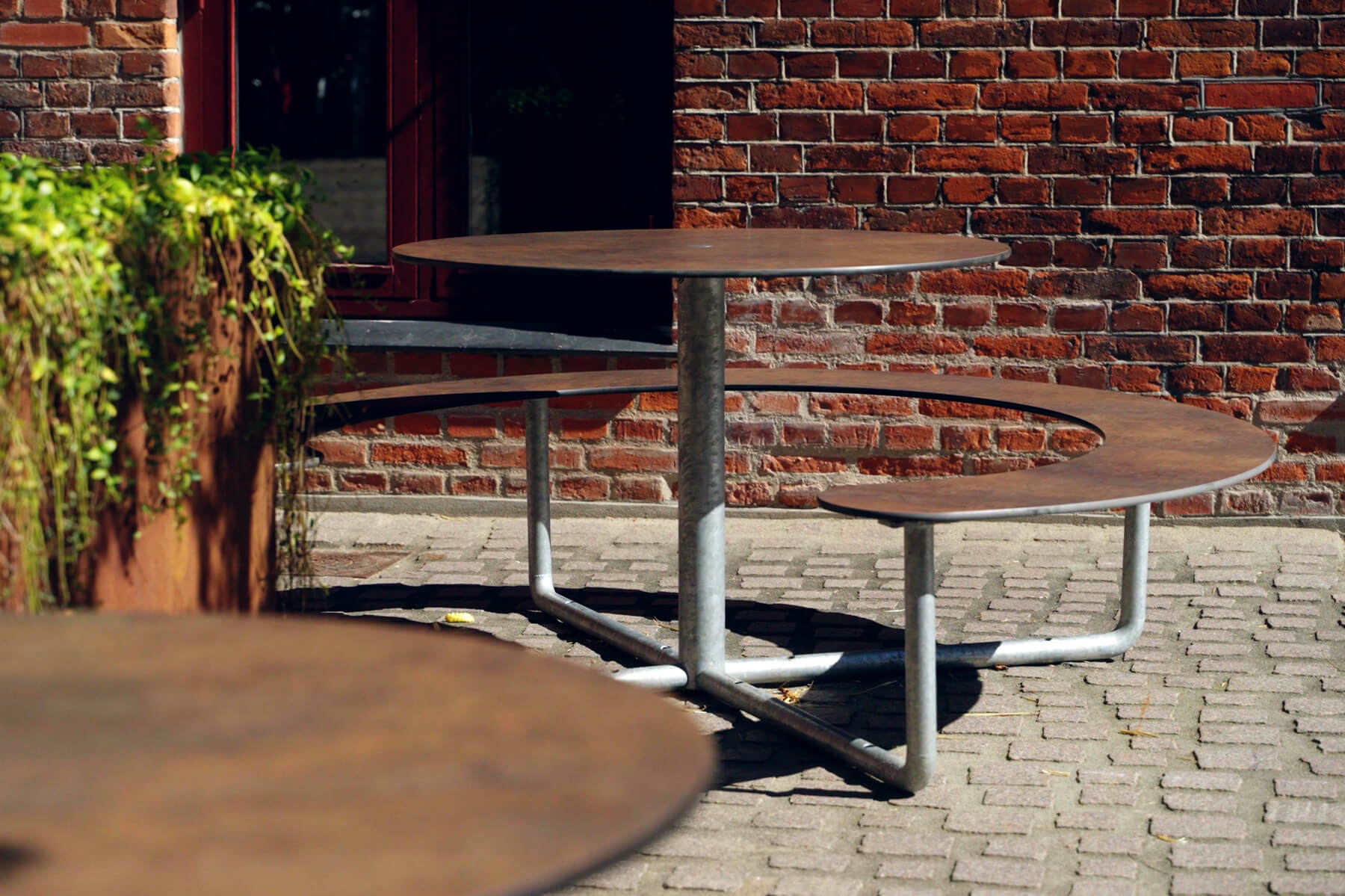 Et nær billede af et brunt Plateau Picnic Round bordbænkesæt på en plads, ved siden af et bed