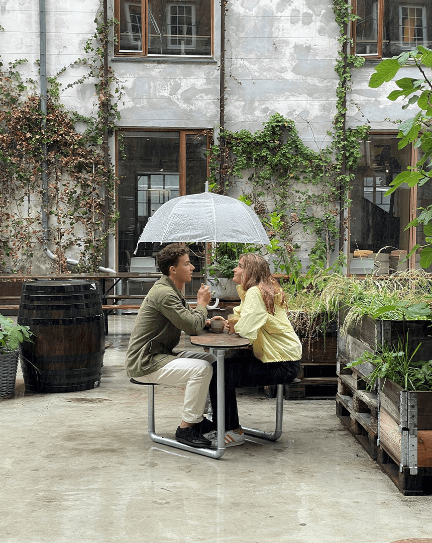 To personer der sidder i læ under paraplyen på et Plateau Picnic Duo bordbænkesæt på en cafe