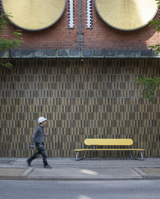En mand der går på et fortov, han passerer en Plateau Bænk i gul farve