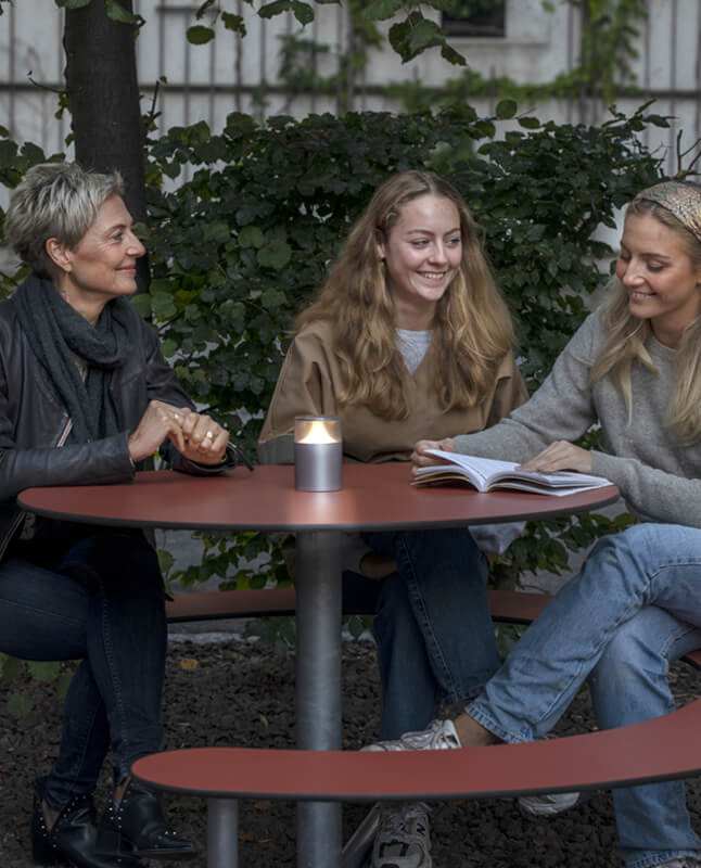 Tre kvinder ved et Plateau Picnic Round bordbænkesæt i en gård om aftenen. Et lys er tændt og den ene læser en bog imens at de to andre kvinder smiler