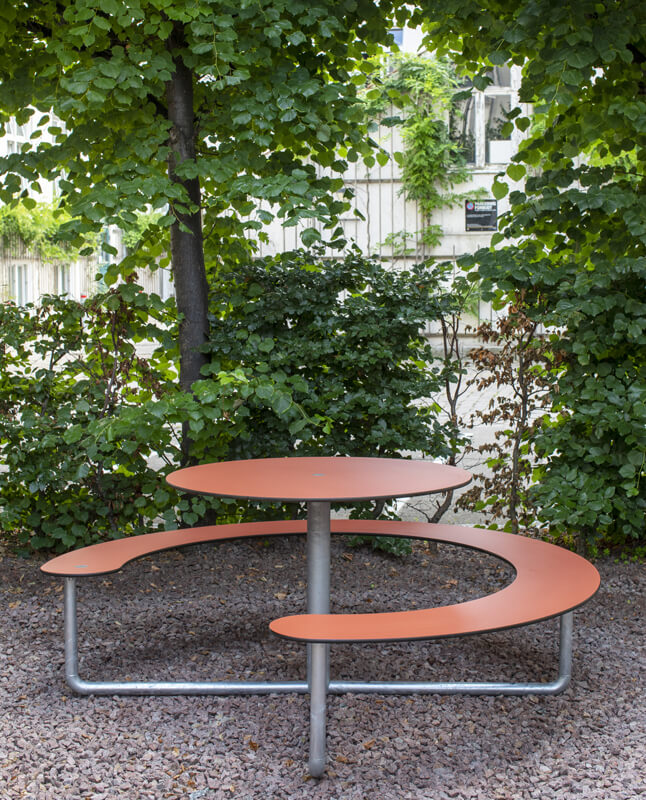 Et Plateau Picnic Round bordbænkesæt der står i en gård, nær træer og buske