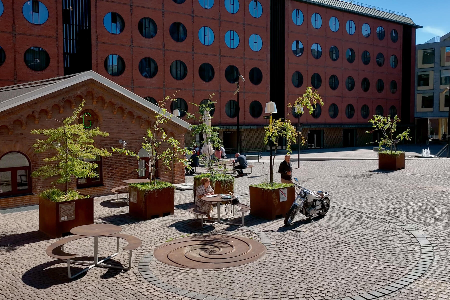 To brune Plateau Picnic Round bordbænkesæt på en plads. Pladsen har træer og bed, og ved siden af ligger Gasolin Grill. En kvinde sidder og nyder udsigten, mens en mand er på vej hen til sin motorcykel.