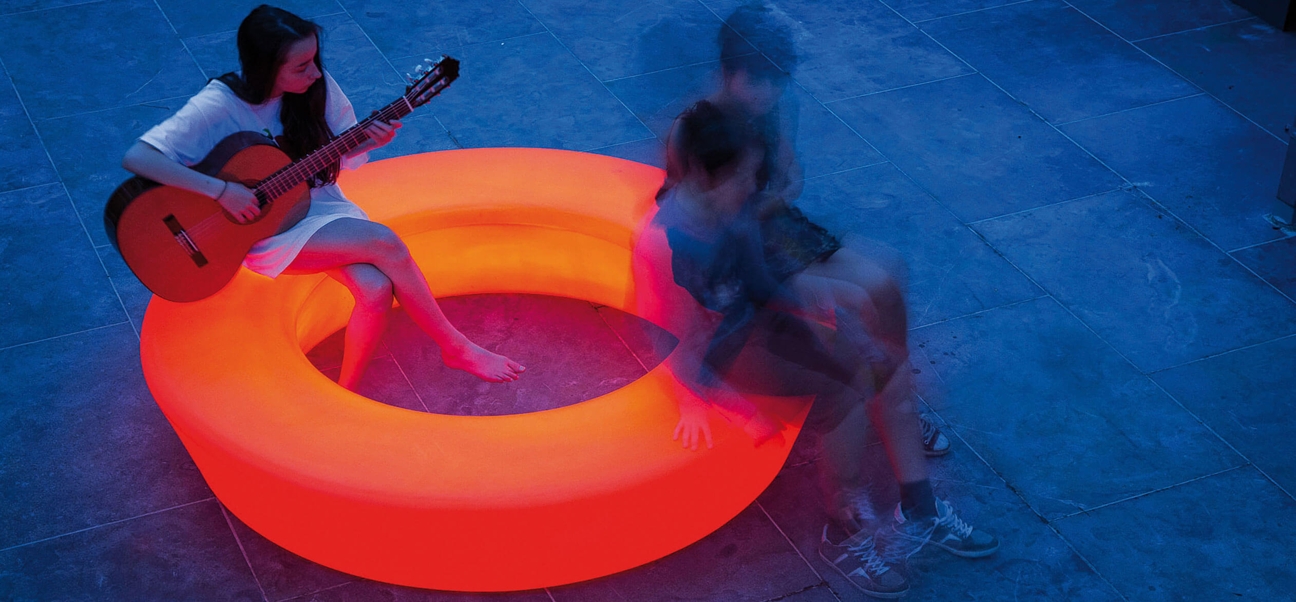 En kvinde der spiller guitar, samt to der snakker på en lysende, rød Loop Light bænk